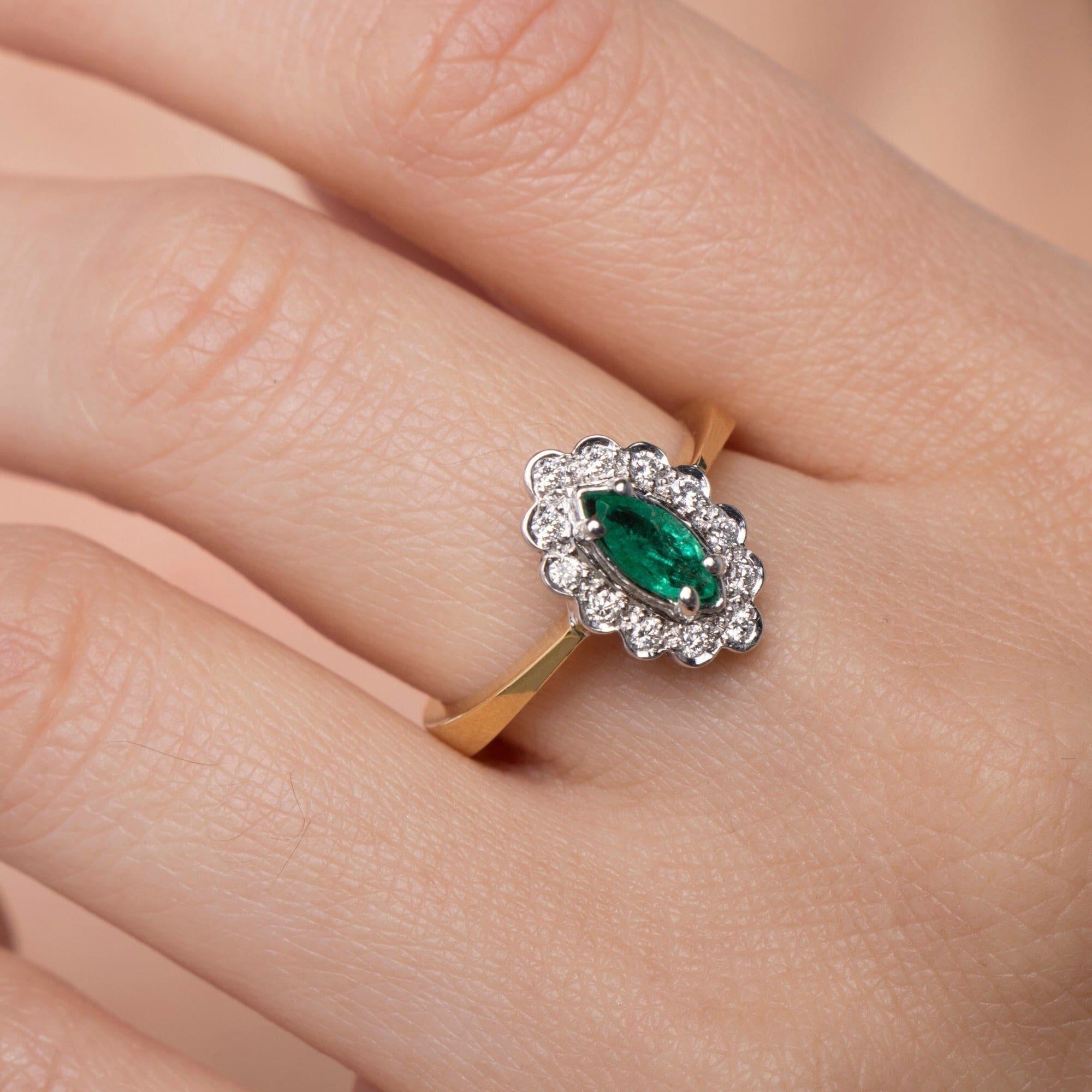 A marquise-cut emerald ring with round-cut natural diamonds set in a 18K yellow gold band, displayed on a person's hand.